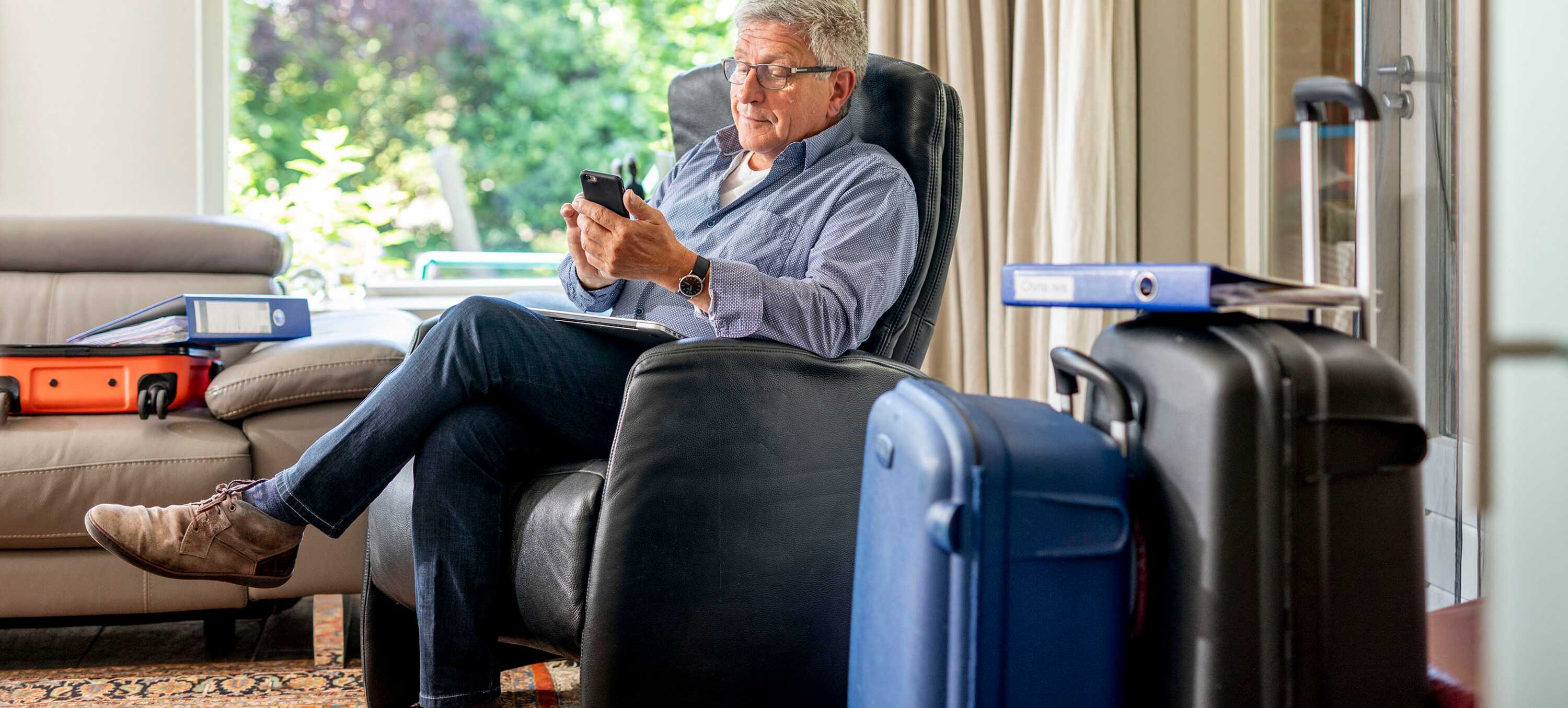 Man op stoel met smartphone. Koffers staan naast hem. 